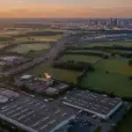 Vue aérienne d'une zone d'activités avec entrepôts et usine, campagne et métropole au loin